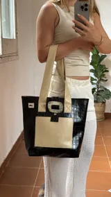 Bolso tote de ecocuero marrón oscuro con textura cocodrilo y un parche frontal de cuero liso marrón claro. Posee asas de hombro de cuero marrón y un broche decorativo blanco en el frente.