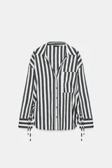 Camisa de cuello solapa con escote pico y manga larga. Presenta un bolsillo delantero de plastrón y bajo con aberturas laterales con cordones ajustables. Cierre frontal con botones.