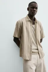 Camisa de calce relajado confeccionada en tejido con mezcla de algodón y lino, con diseño de rayas verticales en tonos beige. Presenta cuello bowling, manga corta, aberturas laterales en el bajo y cierre frontal con botones.