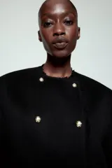 Chaqueta de corte recto confeccionada en mezcla de lana, con cuello redondo y manga larga. Presenta un diseño cruzado con botones metálicos plateados y acabados deshilachados en cuello, puños y ruedo.