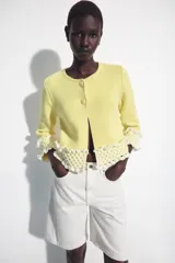 Chaqueta de punto color amarillo claro, con cuello redondo y mangas por debajo del codo. Presenta detalles de acabado en tejido crochet blanco en el ruedo y los puños, con cierre frontal de botones forrados al tono.