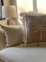 Almohadón cuadrado color marrón claro con bordado de hilo color crema con diseño de rayos de sol. Tiene borde blanco con flecos.