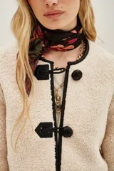 Saco corto de tejido tipo boucle en color crudo, con cierre frontal mediante tres trabas de cuero sintético negro y botones esféricos. Presenta ribetes en contraste negro a lo largo de la abertura frontal y el cuello redondo.