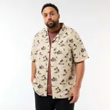 Camisa de manga corta color beige con estampado tropical de palmeras y veleros en tonos oscuros, cuello cubano y un bolsillo aplicado en el pecho.