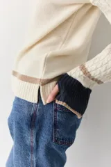 Buzo de punto color crudo con mangas de textura trenzada, detalles en contraste en los puños y franjas horizontales en el ruedo. Presenta cuello redondo y un pequeño bordado en el pecho.
