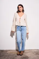 Blusa beige de viscosa y lino, con escote en V, mangas largas acampanadas con detalles bordados tipo broderie, y cierre frontal con lazo para anudar.