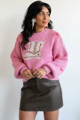 Sweater tejido de punto en color rosado, con diseño de bota estilo western en el frente y detalles de estrellas pequeñas en color rojo. Presenta cuello redondo, mangas largas abullonadas y puños acanalados.