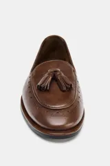 Mocasín de cuero color marrón con detalle de borlas en el empeine y perforaciones decorativas tipo brogue en la puntera y el talón.