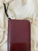 Billetera grande de cuero color borgoña, con cierre metálico y logo de la marca grabado. Incluye moneda colgante plateada con el logo de la marca.