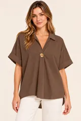 Camisa de algodón color chocolate, de diseño amplio y holgado, con cuello camisero, escote en V pronunciado con un botón decorativo dorado y mangas cortas con vuelta en los puños.