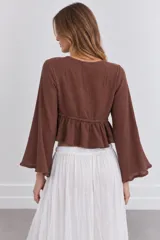 Blusa crop top marrón con escote en V pronunciado, mangas largas acampanadas y cierre de lazo delantero. Tiene detalles de apliques metalizados en el borde del escote y un diseño fruncido en la parte inferior.