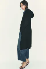 Abrigo de paño negro con capucha, cierre frontal cruzado con botones y cinturón de lazada en el mismo tejido. Presenta manga larga con trabillas en los puños y bolsillos laterales.