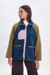Campera de sherpa con diseño color block, combinando cuerpo azul, mangas marrones y detalles en rosa pálido. Presenta cierre frontal con botones a presión, bolsillo superior con cierre y bolsillos laterales con cremallera.