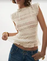 Blusa de tul color crema con textura fruncida y pequeños lunares bordados. Presenta cuello alto, mangas cortas con volados y un corte ajustado al cuerpo con ruedo acampanado.