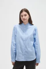 Camisa celeste de manga larga con cuello alto y volados, detalles de frunces en hombros y puños, y cierre frontal con botones.