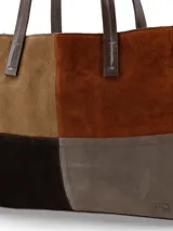 Cartera tipo tote bag confeccionada en gamuza con diseño de paneles en tonos marrón, chocolate y tostado. Cuenta con doble asa de hombro en cuero liso marrón oscuro y detalles de costuras visibles.