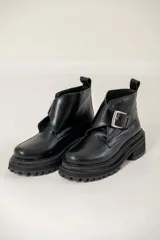 Bota corta de cuero negro con suela track dentada y taco cuadrado. Presenta una tira decorativa en el empeine con una hebilla metálica plateada y tirador en la parte trasera.