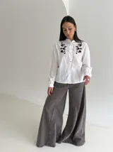 Camisa blanca de manga larga con cuello mao, cierre frontal de botones y detalle de volado. Presenta bordados florales en color marrón chocolate en la parte superior delantera.