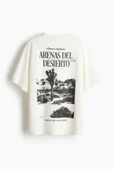 Remera oversized de punto suave de algodón, color blanco, con cuello redondo acanalado, hombros caídos y diseño estampado en el frente con la frase 'Arenas del Desierto'.