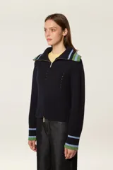 Cárdigan de punto azul marino con cierre frontal de cremallera. Presenta cuello tipo camionero y puños con detalles de rayas en tonos verde y celeste. El tejido tiene una textura acanalada con detalles calados decorativos.