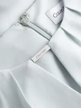 Blusa celeste de poliéster reciclado con cuello redondo, mangas cortas y cierre con cremallera en la espalda. Presenta una abertura en la parte delantera con una barra metálica decorativa.