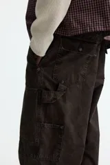Pantalón estilo carpenter de corte holgado, confeccionado en tejido de algodón color marrón oscuro. Presenta tiro caído, bolsillos laterales de parche, trabilla funcional en una pierna y costuras reforzadas visibles sobre y debajo de las rodillas.