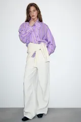Camisa de corte oversize con diseño de rayas verticales en tonos violeta, blanco y bordó. Presenta cuello subido cruzado con cierre de botones frontal, mangas largas con puños y bajo asimétrico.