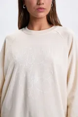 Buzo deportivo de corte oversize en color crudo, con cuello redondo y mangas largas. Presenta un diseño bordado en el frente con la inscripción Saint Tropez y un motivo floral central.