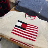 Sweater de tejido de punto color beige con cuello redondo y detalle de ribete rojo. Presenta un diseño frontal grande con el logo de la bandera de Tommy Hilfiger en colores blanco, rojo y negro.