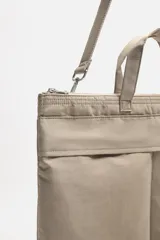 Bolso tipo shopper de tejido técnico color beige, con dos asas de mano y una correa de hombro ajustable y extraíble. Presenta un compartimento principal con cierre de cremallera, un bolsillo interior con cremallera y dos bolsillos frontales con cierre de botón a presión.