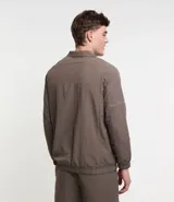 Campera deportiva color marrón, con cuello alto, cierre frontal con cremallera, manga larga, bolsillos laterales y detalles de etiqueta aplicada y rayas.