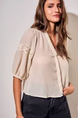 Blusa de color arena con cuello en V, mangas tres cuartos abullonadas y detalles de encaje bordado en hombros y mangas. Presenta un diseño ligero y delicado con cierre frontal.