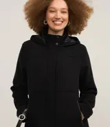 Saco largo de polivelour color negro, con cuello alto, capucha y cierre frontal de cremallera y botones. Presenta bolsillos laterales con cierre y forro interno.