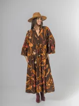Vestido largo de manga larga, corte oversized y cuello camisero con abertura en V y botones. Presenta un estampado botánico en tonos tierra sobre fondo marrón oscuro, con volados en el ruedo.