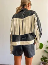 Campera negra de cuero sintético con cierre frontal, detalles en blanco en cuello, puños y ruedo, y flecos blancos en las mangas.