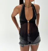 Top halter negro con escote profundo en V, confeccionado con tela opaca en la parte superior y tela transparente en la parte inferior. Se anuda al frente con un broche metálico plateado, del cual cuelgan tiras largas de cuentas plateadas con dijes colgantes.