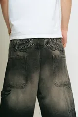 Bermuda de jean estilo carpintero, corte baggy, color negro lavado con efecto degradado. Presenta bordados en la parte trasera con tipografía gótica que dice "Lucca Bossio". Incluye bolsillos grandes y bolsillo lateral de carpintero.