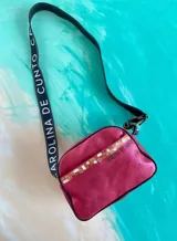 Bandolera Kaira de la línea Ottenta de Carolina De Cunto, en color fucsia metalizado. Cuenta con un diseño compacto, bolsillo exterior con broche imantado y detalle pintado a mano, cierre superior y una correa larga regulable con el logo de la marca estampado.