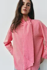 Camisa de rayas rosadas y blancas, con cuello subido decorado con un lazo integrado, manga larga con puños abotonados y cierre frontal oculto.
