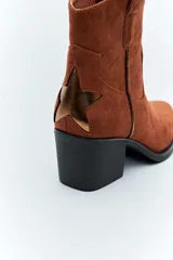 Bota corta de gamuza color negro, estilo texana, con taco cuadrado y puntera ligeramente perfilada. Presenta costuras decorativas en el empeine y tiradores laterales.