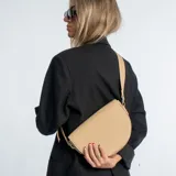 Cartera bandolera de ecocuero color beige, con forma de media luna y correa al hombro ajustable.