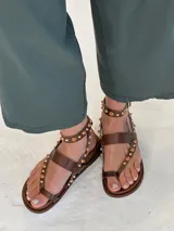 Sandalias planas de cuero marrón, con diseño de tira al dedo (toe-post), tira horizontal en el empeine y tira ajustable en el tobillo. Tienen apliques de tachas metálicas doradas y piedras naranjas a lo largo de las correas.