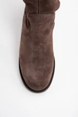 Bota de caña alta confeccionada en gamuza color marrón, con diseño de caña holgada y suela plana con pequeño taco cuadrado.