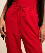 Pantalón de crepé rojo, modelo estilo pijama, de corte fluido, con cintura elástica y lazo de ajuste con terminales metálicas. Cuenta con bolsillos laterales y cierre frontal con cremallera y botón.