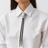 Collar bolo de cuero trenzado negro con un aplique central de metal plateado con detalles grabados y una piedra clara en el centro, y punteras metálicas decoradas.