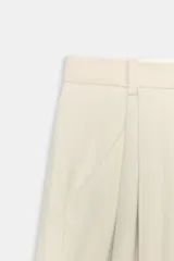 Pantalón de tiro medio con pierna ancha, confeccionado en una mezcla de lino y algodón con tejido en espiga. Presenta pliegues frontales, bolsillos laterales y cierre frontal con cremallera, botón y ganchos metálicos.