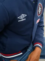 Campera bomber azul marino estilo retro, con cierre frontal de botones a presión, cuello, puños y ruedo con tejido acanalado tricolor. Presenta el logo de Umbro y el escudo del Club Nacional de Football bordados en el pecho, junto con el año 1899 y parches decorativos en las mangas.