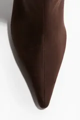 Botas altas de color marrón oscuro, con caña recta y punta fina. Cuentan con un taco fino y alto, y parecen ser de material tipo gamuza o tejido.