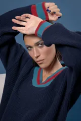 Buzo de punto en color azul marino con escote en V, puños y cuello con detalles en franjas de color turquesa y rojo. Presenta un diseño holgado de manga larga.