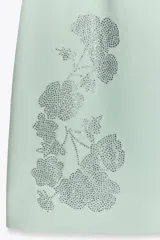 Vestido corto color verde agua, entallado, con escote recto y hombros descubiertos. Presenta apliques de strass con forma de flores. Cierre trasero con cremallera oculta.